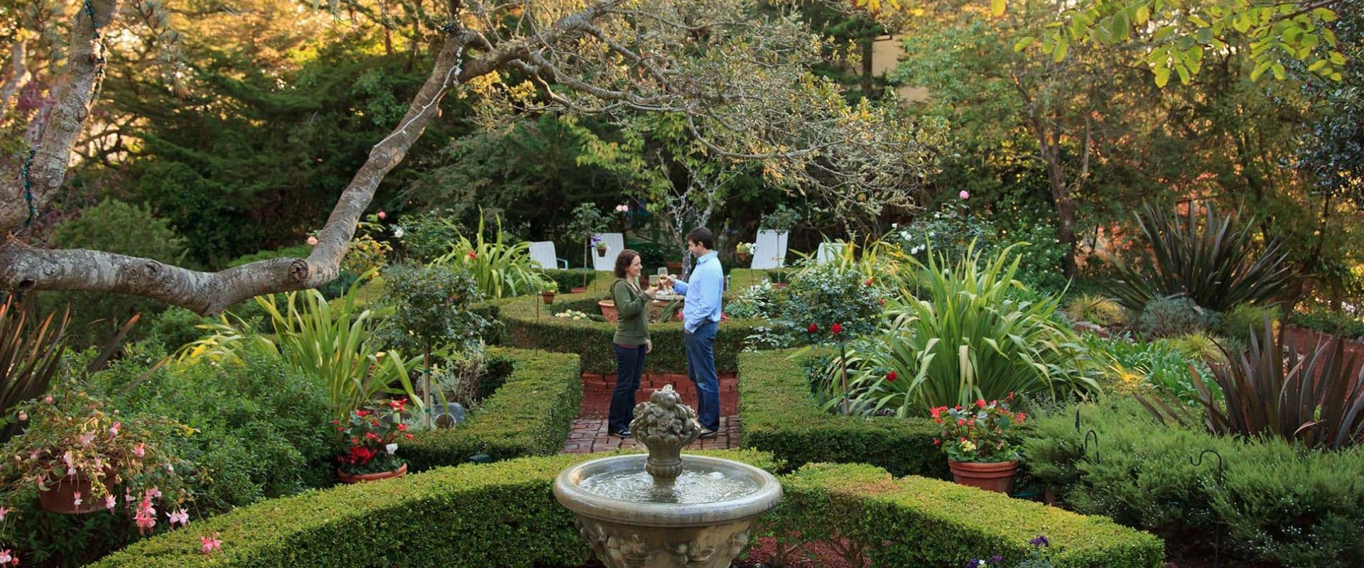 A couple converses in a lush garden with a fountain and vibrant flowers.