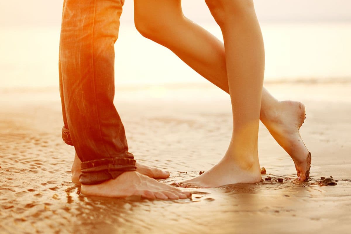 Two people stand barefoot on a beach at sunset, their legs intertwined.