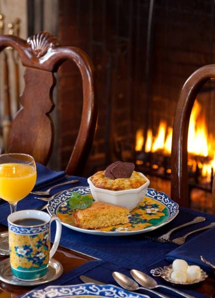 A cozy breakfast setting featuring a plate of cornbread, a bowl of baked dish, a glass of orange juice, and a cup of coffee, all by a warm fireplace.