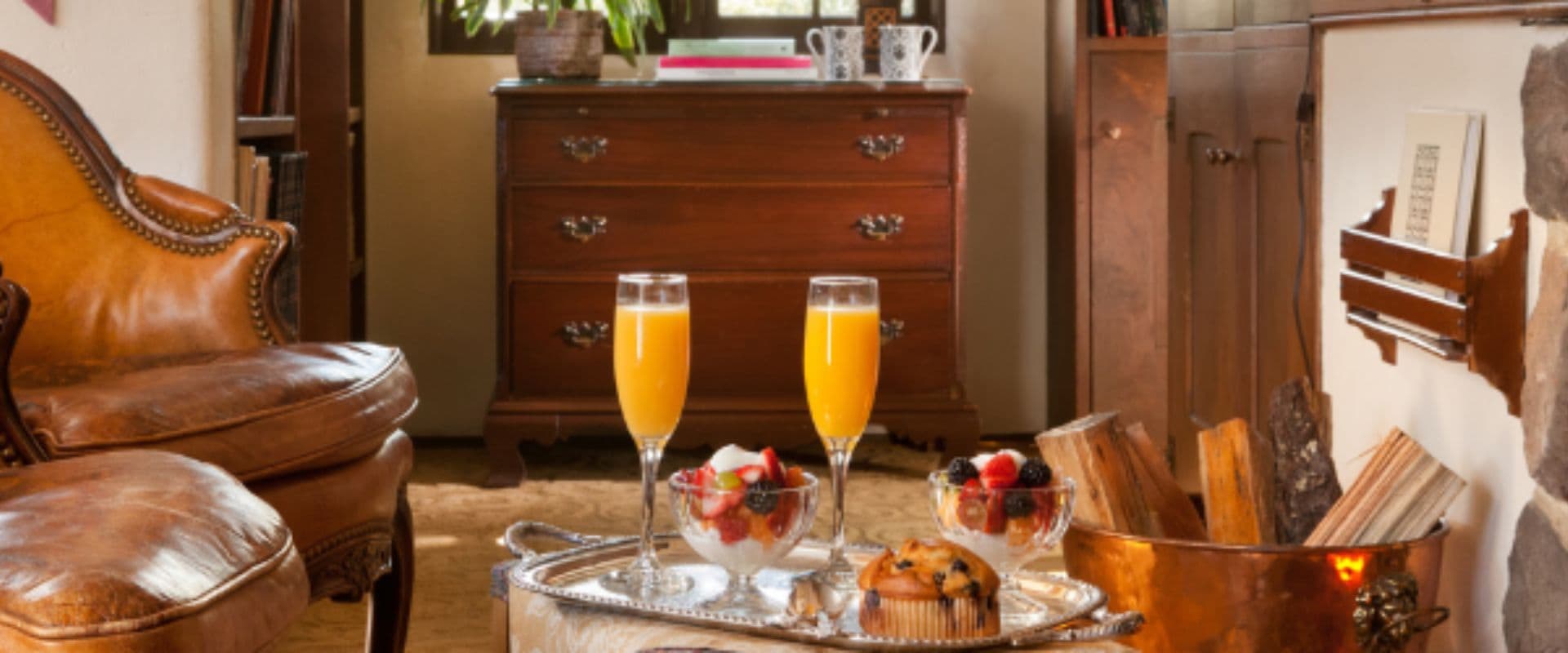 A cozy living room scene featuring two glasses of orange juice, fruit bowls, and a muffin on a silver tray beside a leather armchair.