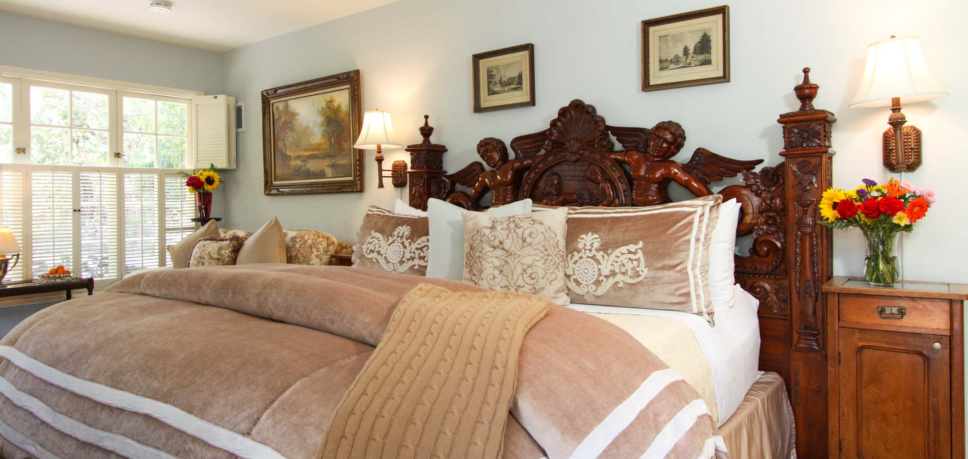 A cozy bedroom featuring an ornate wooden bed, plush bedding, and bright floral arrangements.