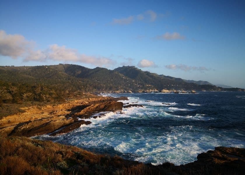 A rugged coastline with crashing waves and rolling hills under a blue sky.
