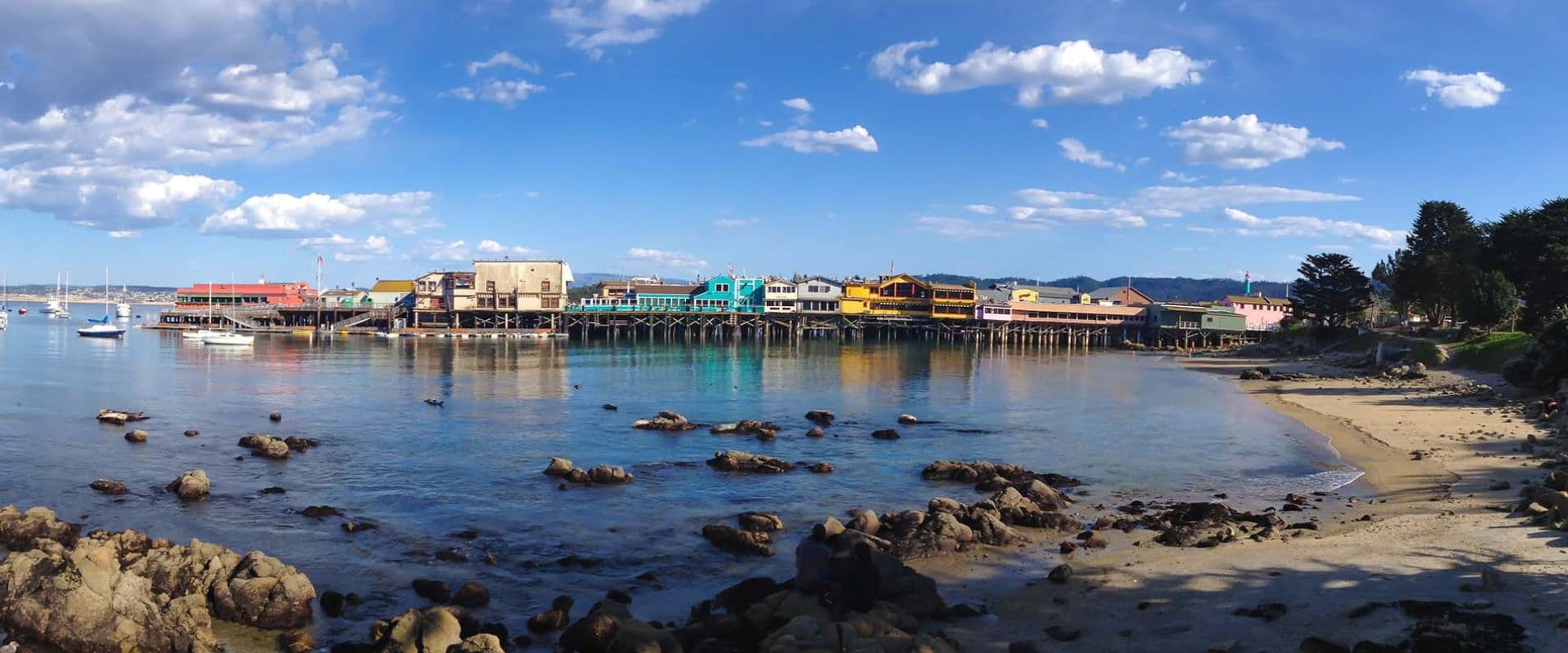 Colorful buildings line the shore of a calm bay under a blue sky with fluffy clouds.