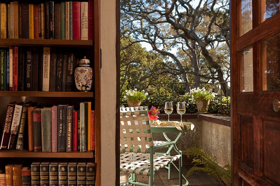 A bookshelf beside an open door reveals a charming patio with flowers and two glasses on a table.