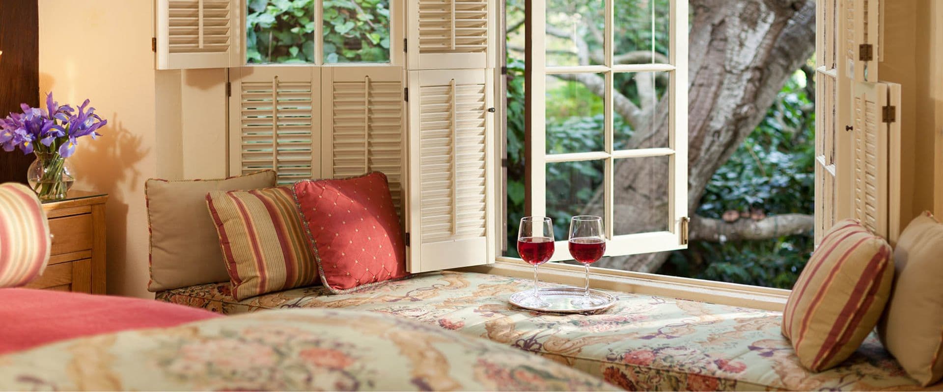 A cozy window seat with decorative cushions and two glasses of red wine overlooking a green garden.
