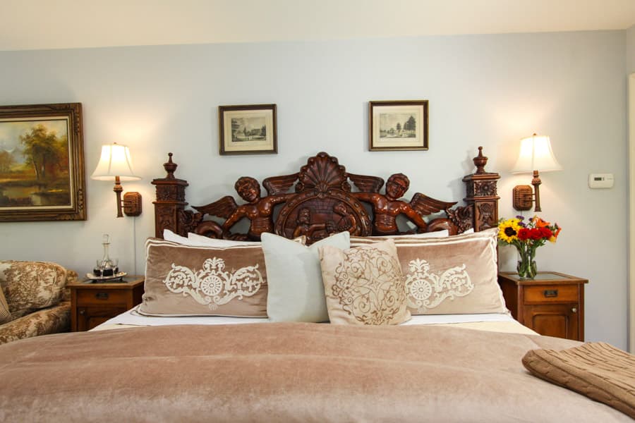 A cozy bedroom featuring a carved wooden headboard, plush bedding, and decorative pillows, complemented by framed art and a bouquet of flowers.