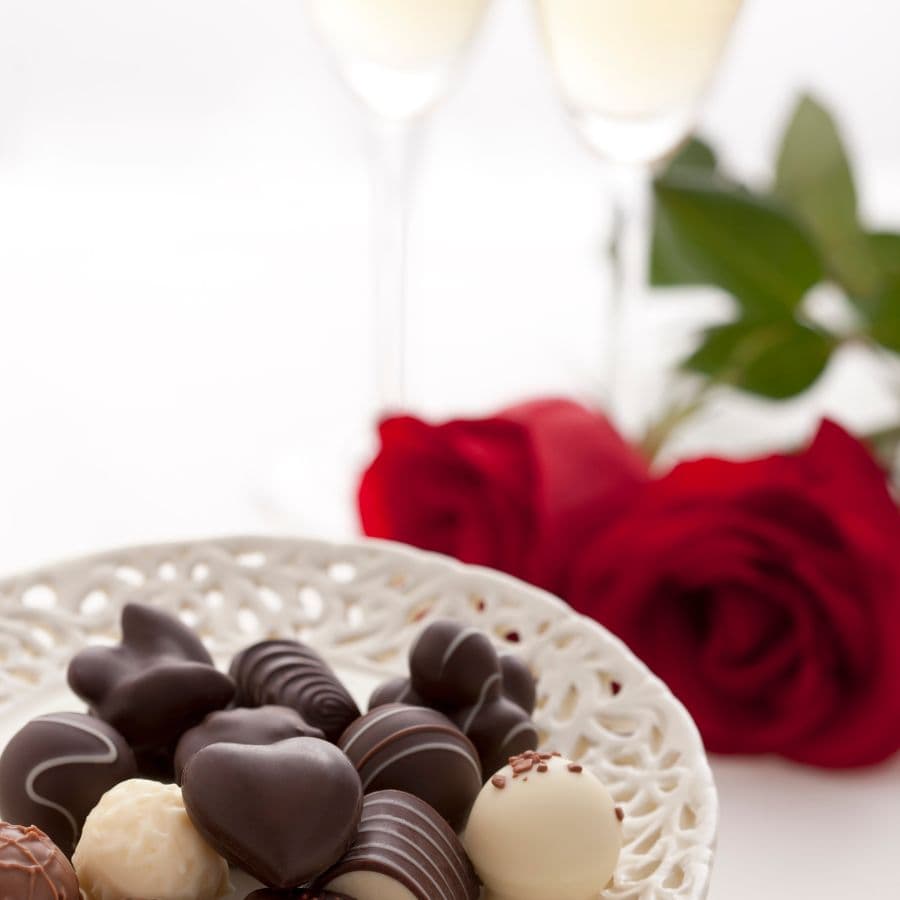 A plate of assorted chocolates with red roses and flutes of champagne in the background.