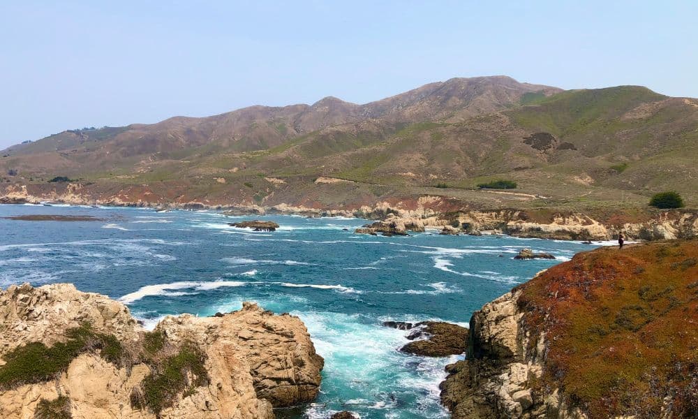 Rugged Garrapata State Park coastline with rocky cliffs, crashing waves, and rolling coastal hills beneath a cloudy sky.