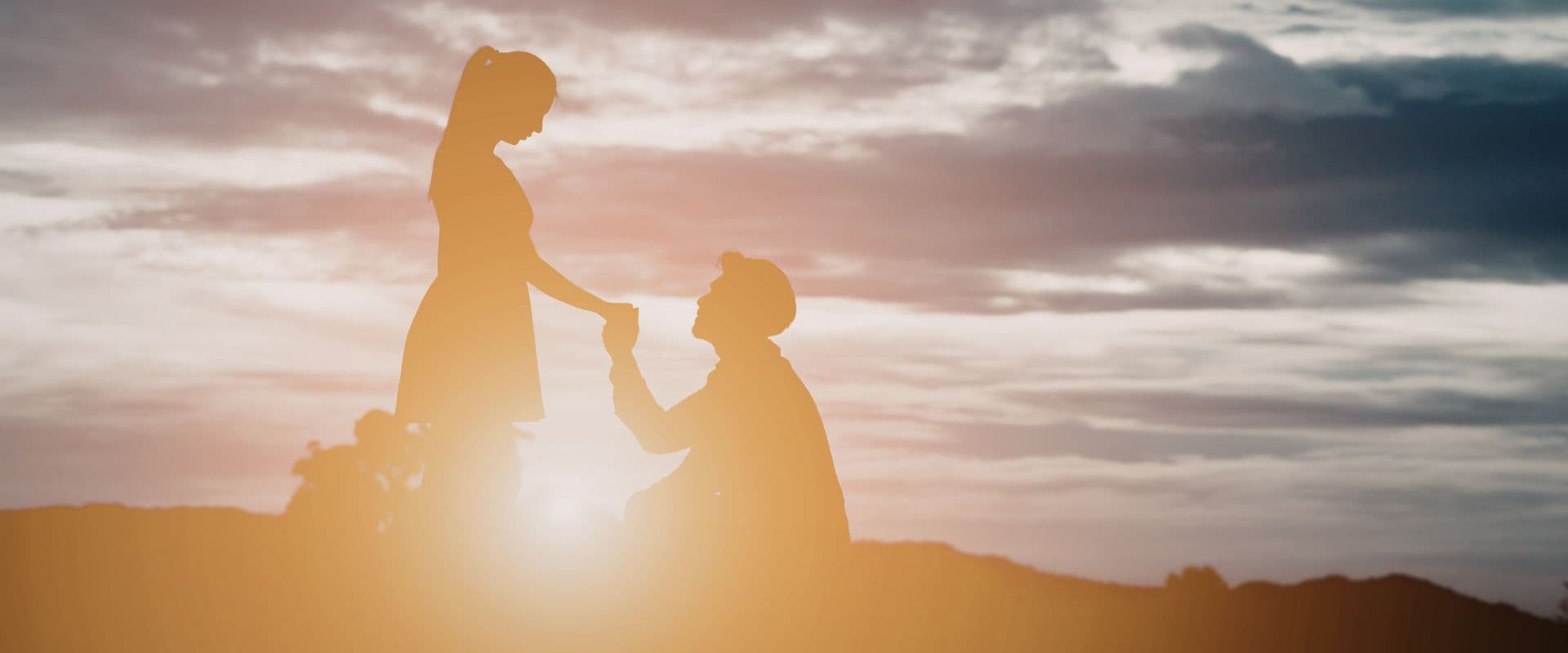 A silhouetted couple shares a romantic moment during sunset, with one partner kneeling and holding the other's hand.