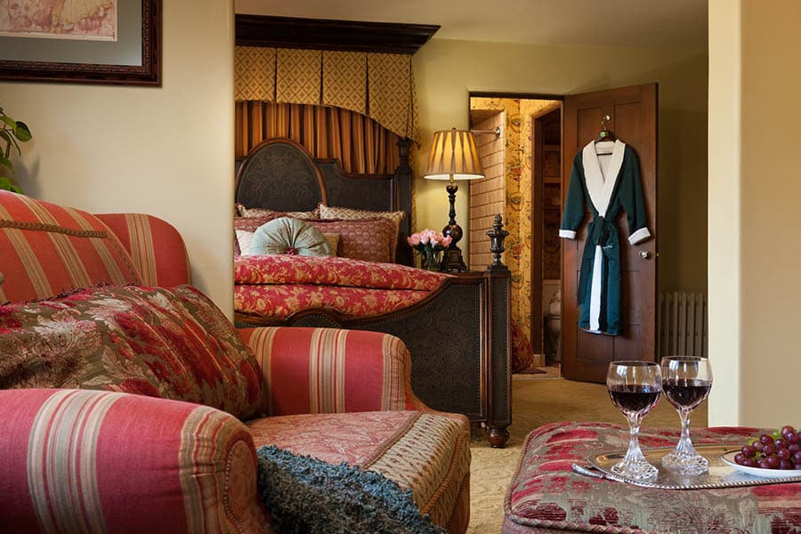A cozy bedroom featuring a plush bed, striped armchair, and glasses of wine on an ornate table.