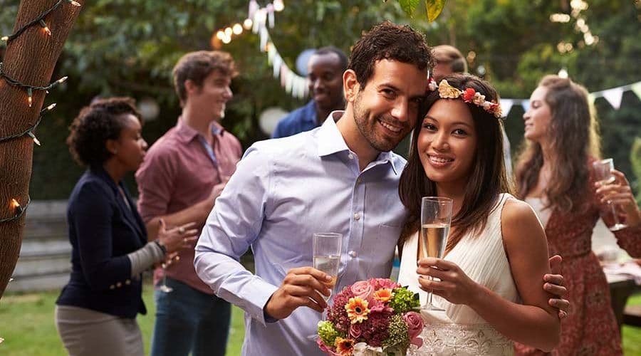 A smiling couple holds glasses of champagne at a lively outdoor celebration surrounded by friends.