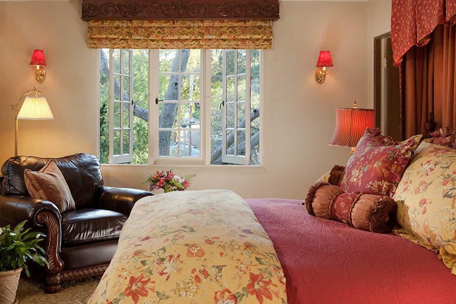 Cozy bedroom with floral bedding, a leather chair, and windows overlooking greenery.