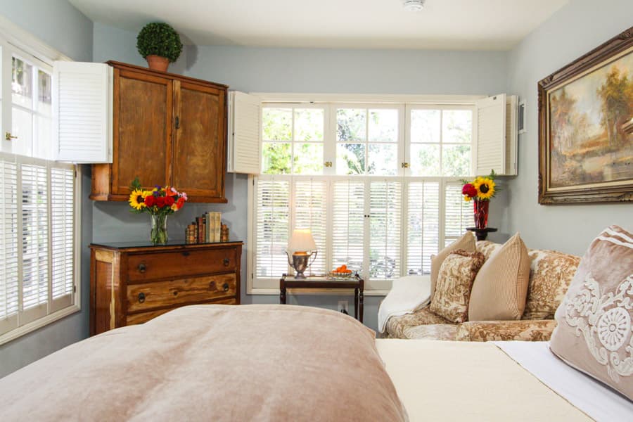 A cozy bedroom with soft bedding, a wooden chest, flower arrangements, and large windows letting in natural light.