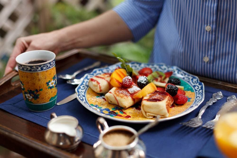 A hand reaches for a colorful plate of fruit and sweet rolls beside a cup of coffee on a blue tablecloth.