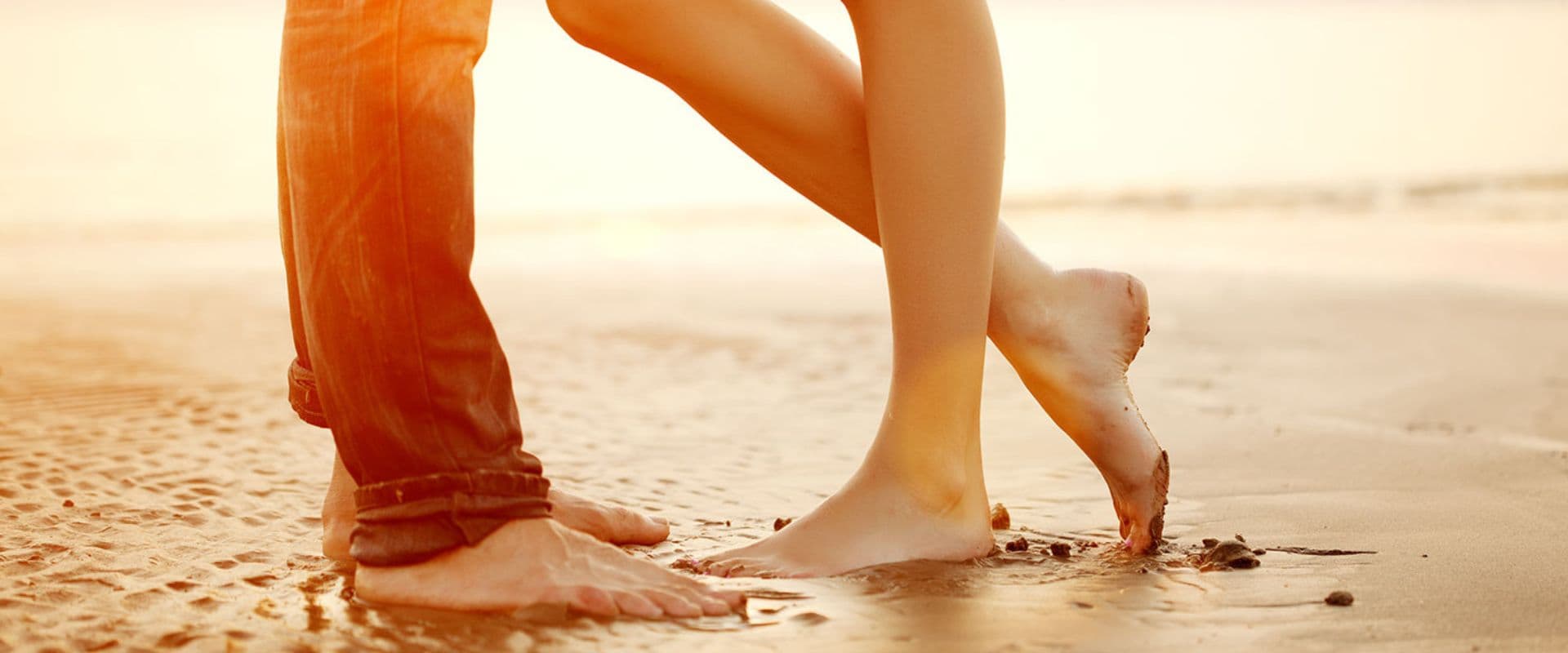 A couple stands barefoot on a beach, gently touching each other's legs.