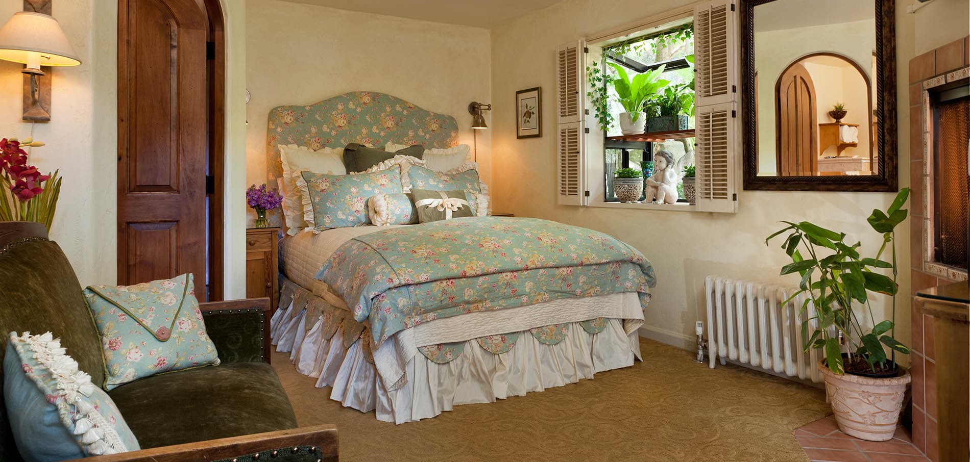 A cozy bedroom featuring a floral-patterned bedspread, a comfortable seating area, and natural light streaming through a window with plants.