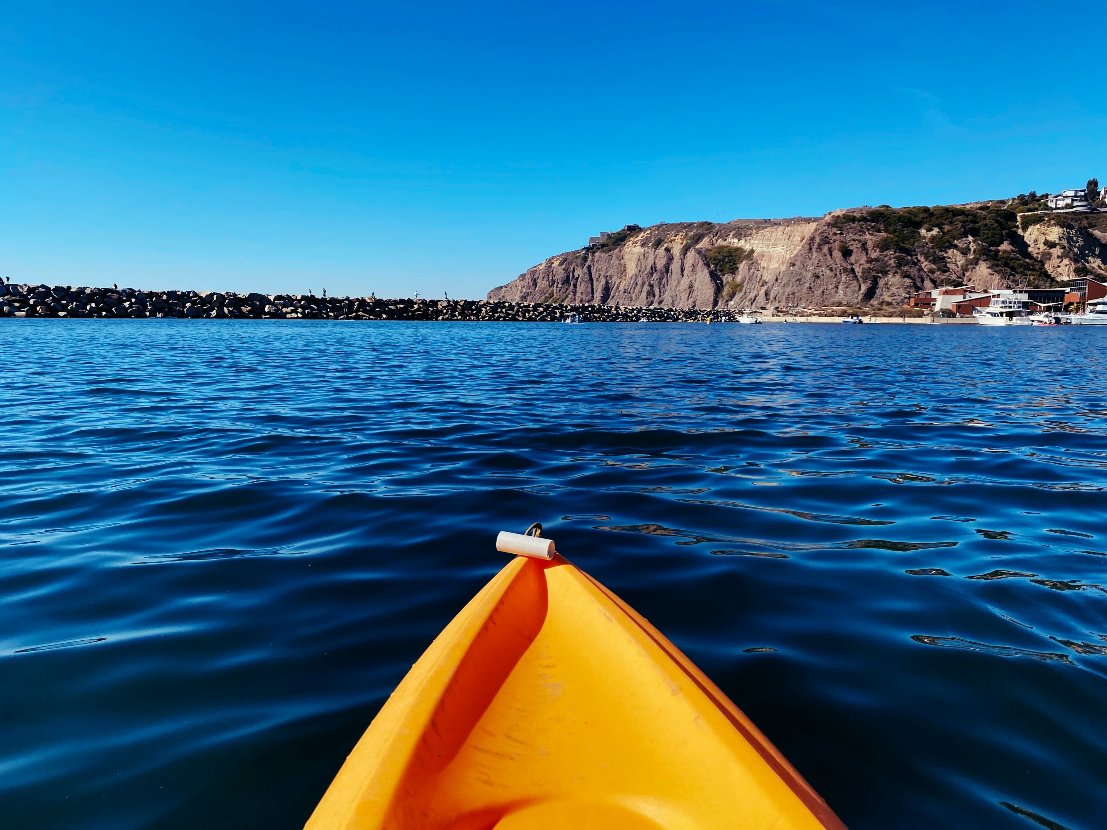 A view from the front of an orange kayak on calm water, facing a rocky cliff under a clear blue sky.