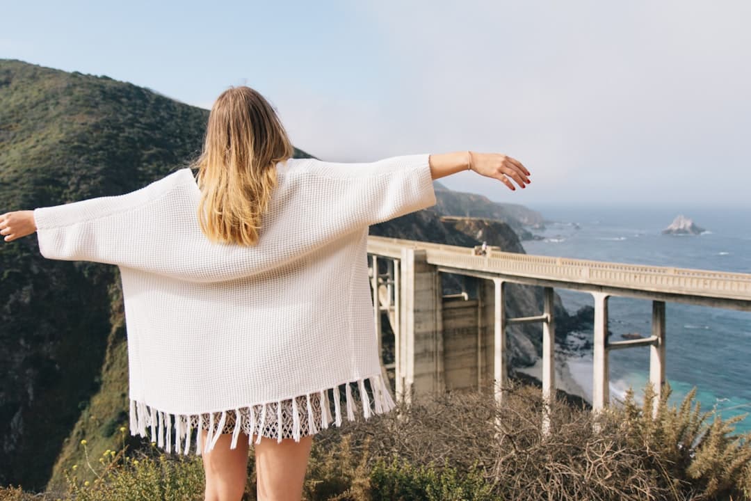A woman in a white sweater stands on a hillside, arms outstretched, overlooking a coastal highway and ocean.