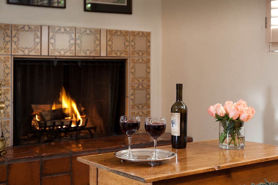 A cozy living room with a roaring fireplace, two glasses of red wine on a wooden table, and a vase of pink roses.