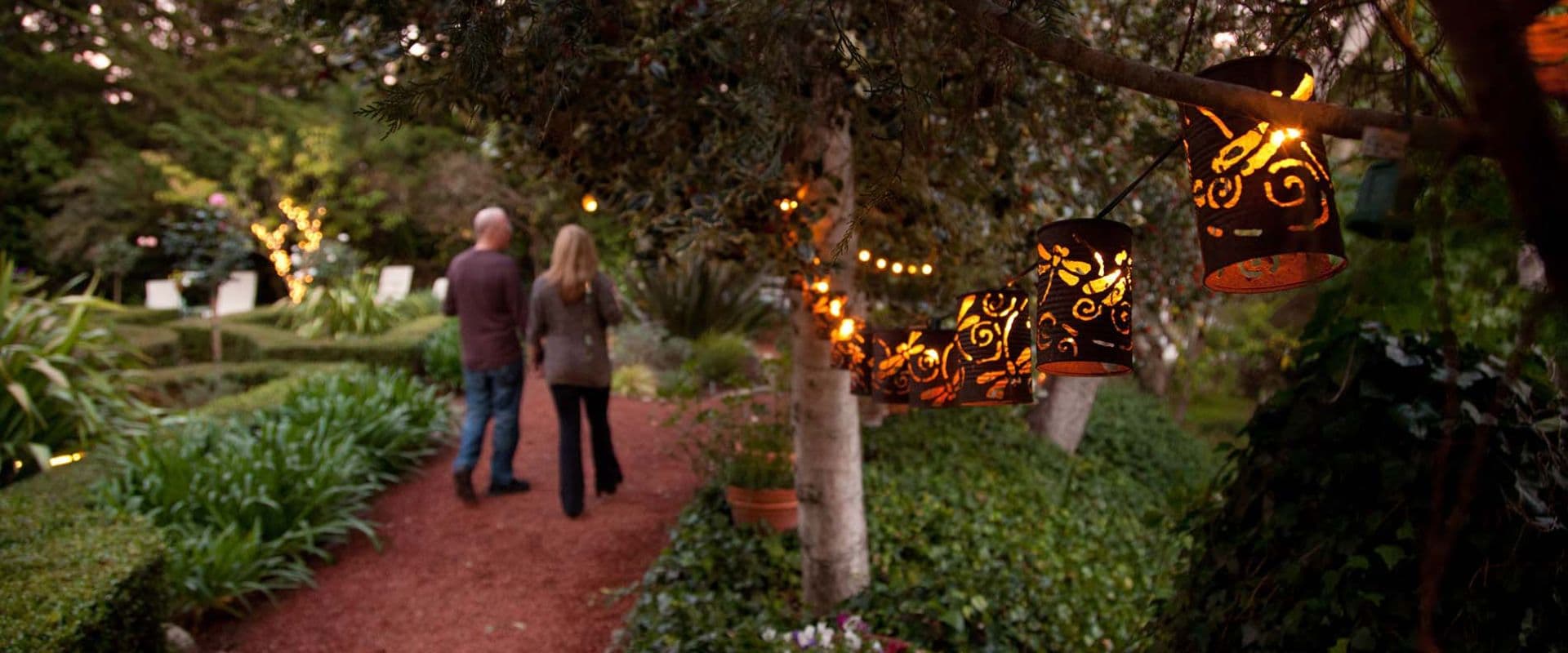 A couple walks along a path in a garden illuminated by decorative lanterns.