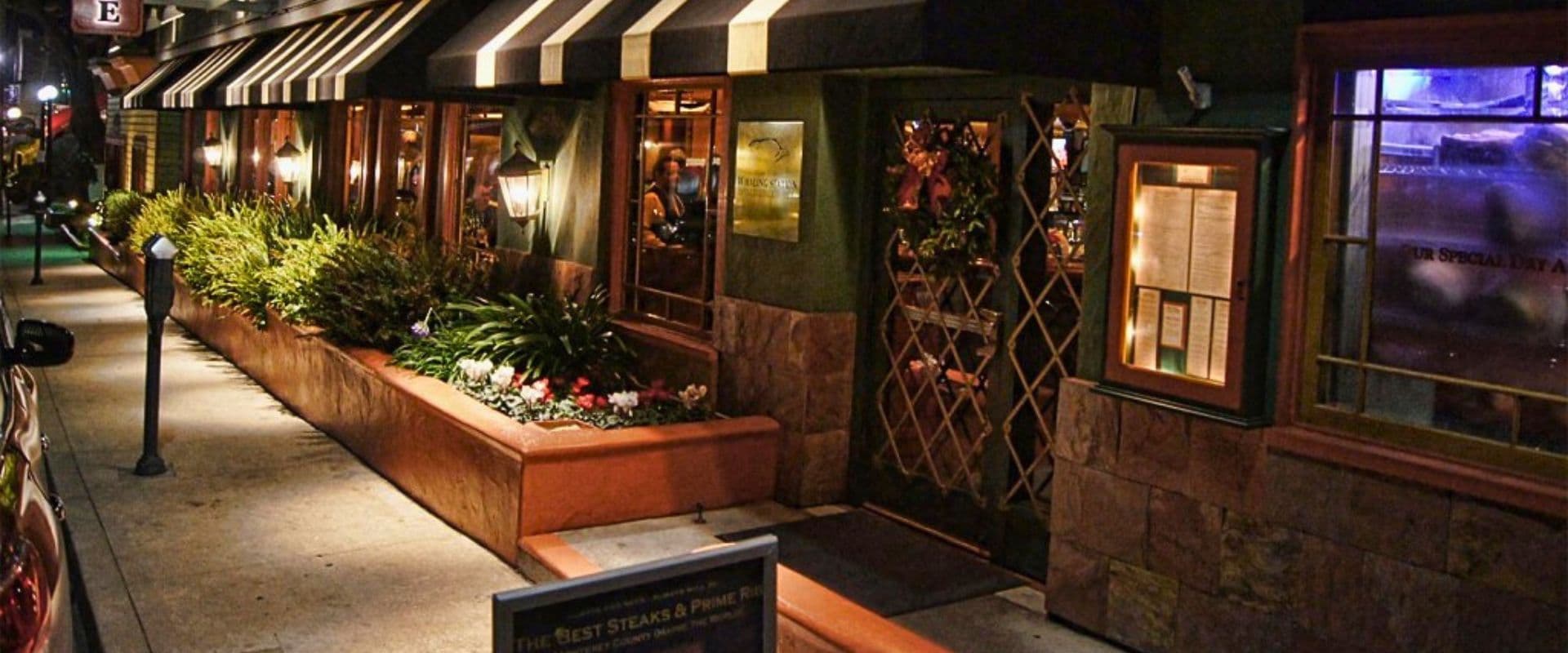 A warmly lit restaurant exterior featuring large windows and lush greenery at night.