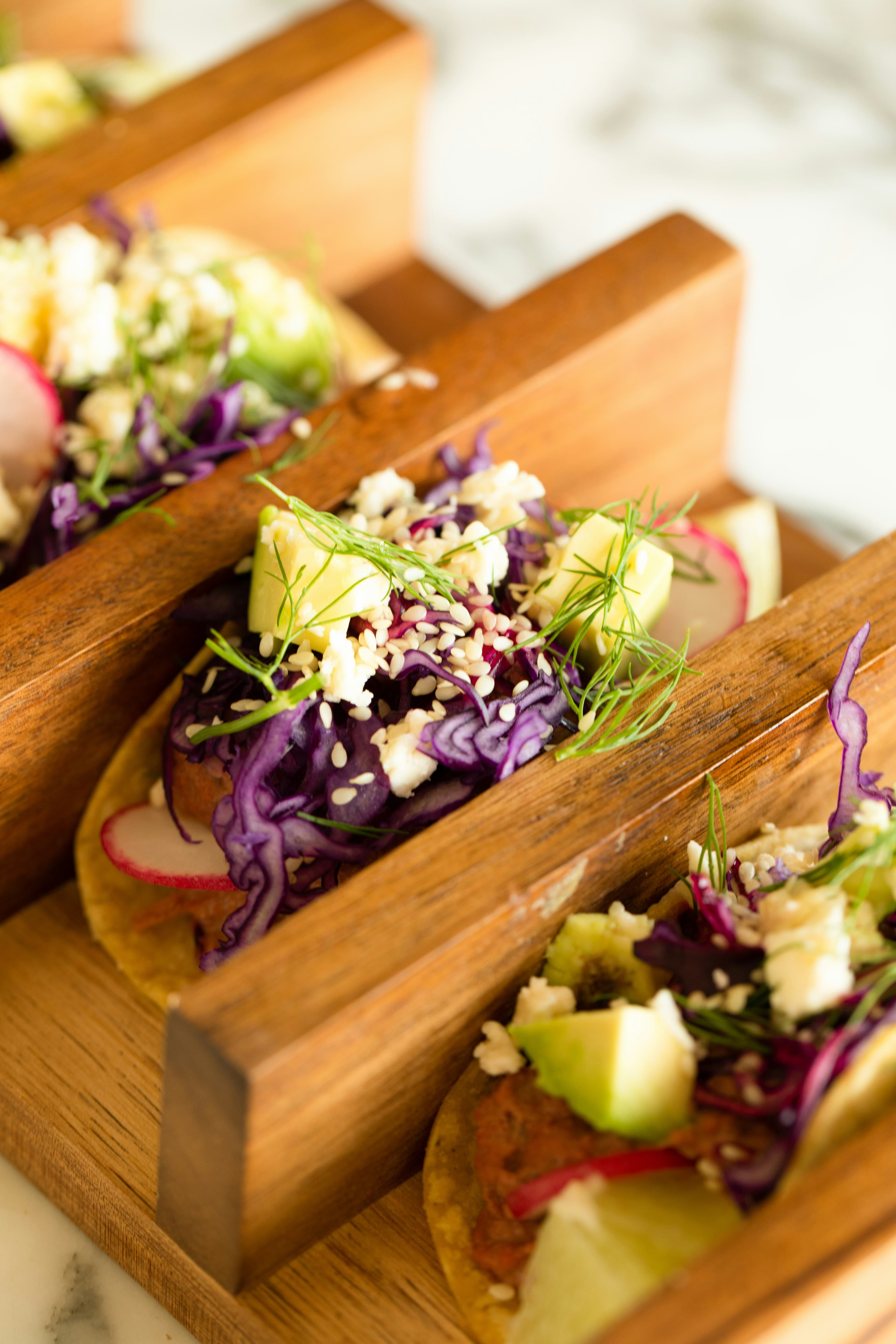 A close-up of colorful tacos topped with purple cabbage, radishes, and herbs served in a wooden holder.