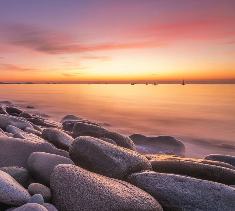 A serene sunset over calm waters, with smooth stones lining the shore.