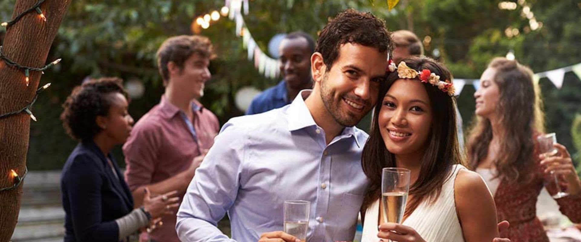 A smiling couple holds drinks at a lively outdoor party surrounded by friends.