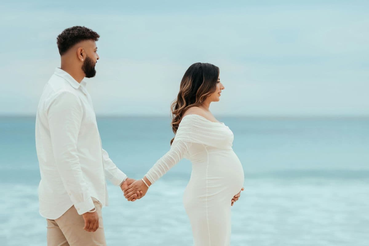 A couple holds hands on the beach, with the woman in a white dress showing her baby bump.