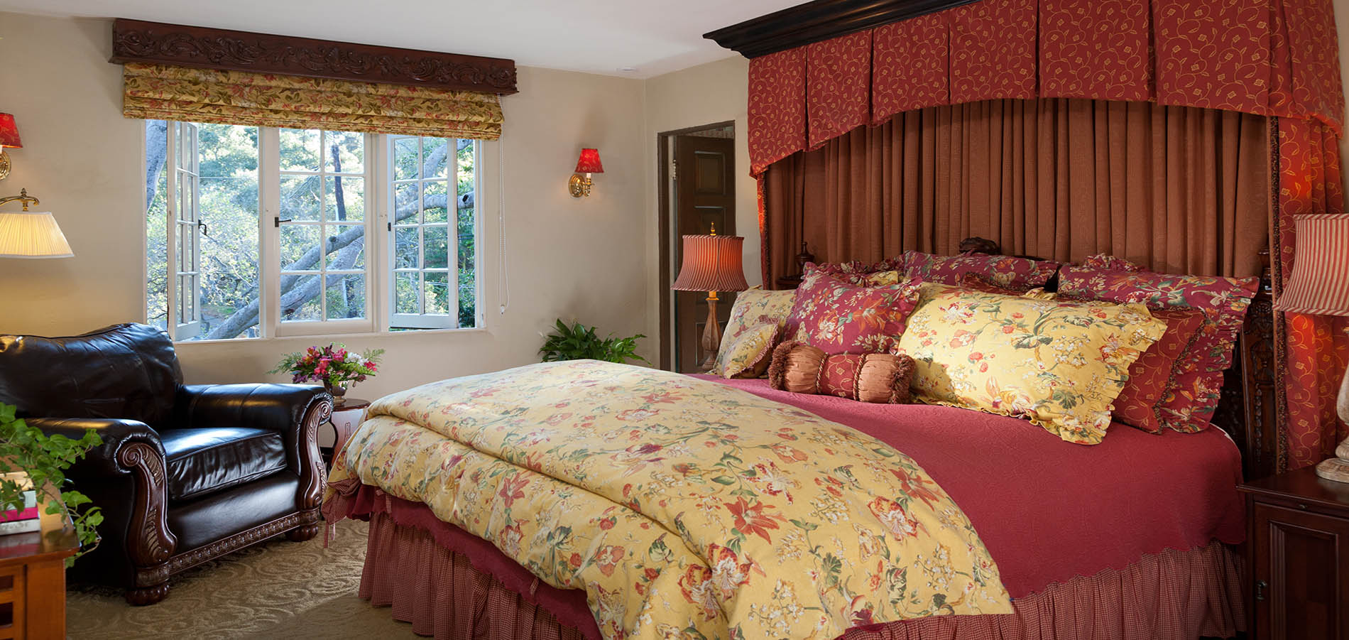 A cozy bedroom featuring a large bed with floral bedding, a leather armchair, and natural light from a window.