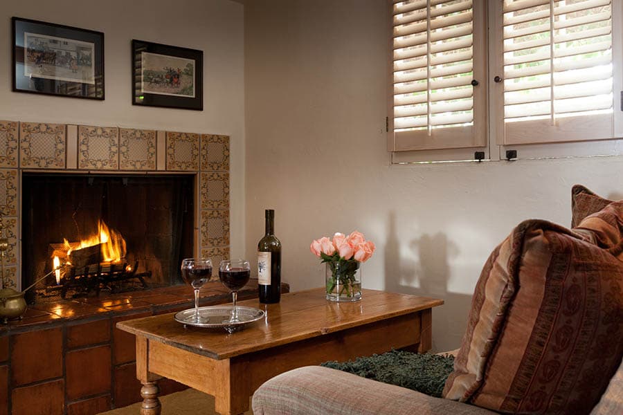 A cozy living room featuring a fireplace, a wooden coffee table with wine glasses, and a vase of pink roses.