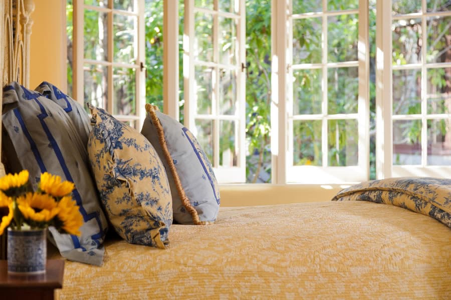 A cozy bedroom with a yellow bedspread, decorative blue pillows, and sunlight streaming through multiple windows.