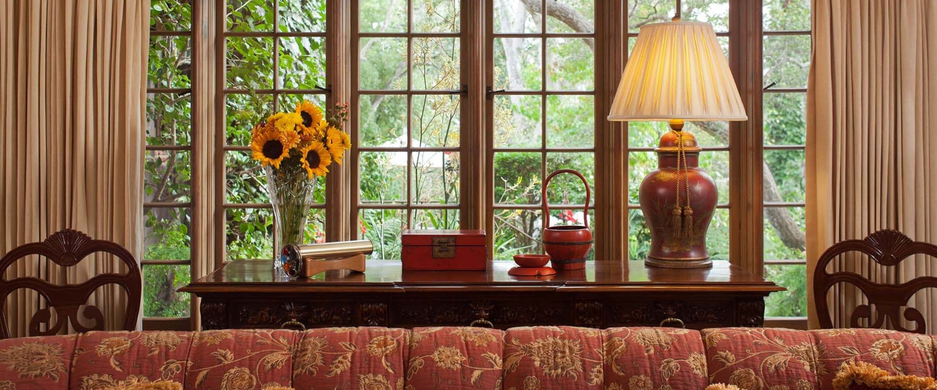 A cozy living room scene featuring a floral arrangement, a lamp, and a wooden table with various decor items, viewed through large windows.