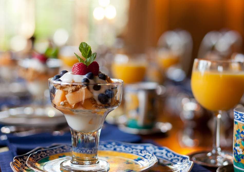 A glass dessert filled with yogurt, fruit, and granola, garnished with mint, against a backdrop of a beautifully set table.