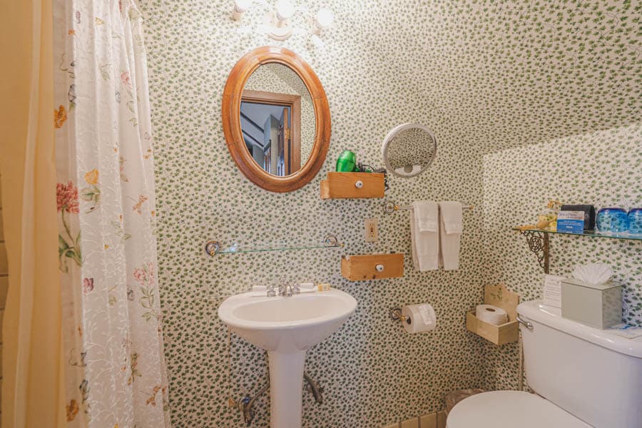 A quaint bathroom featuring floral wallpaper, a circular mirror, and a pedestal sink.