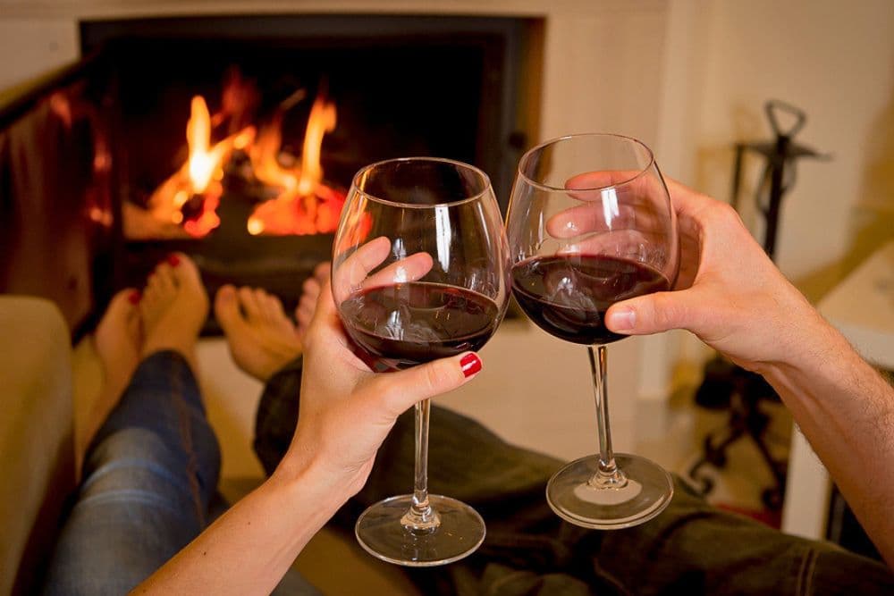 Two hands clink wine glasses in front of a cozy fireplace.