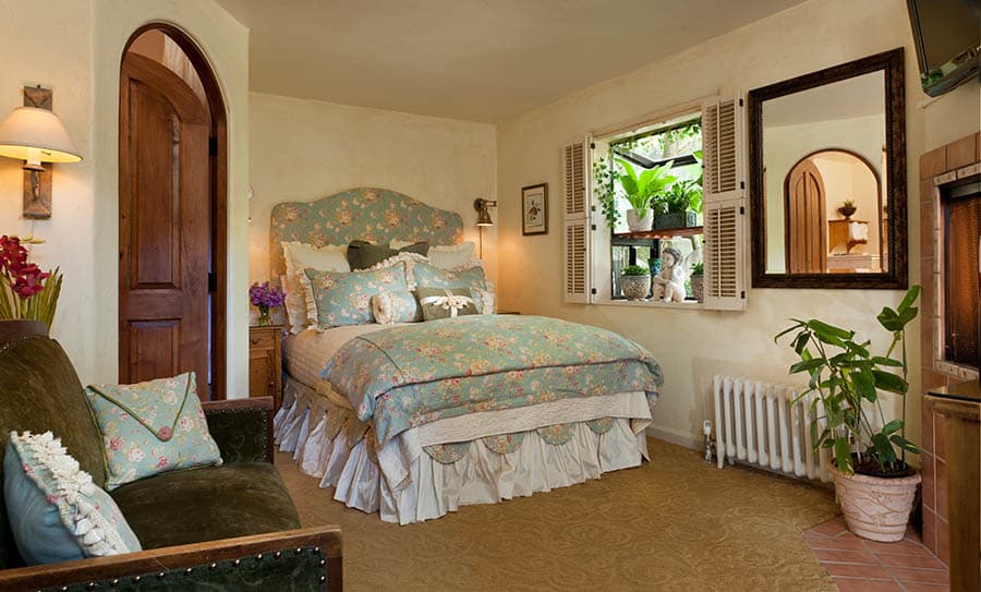 A cozy bedroom featuring a flower-patterned bedspread, plush seating, and natural light from a window with plants.
