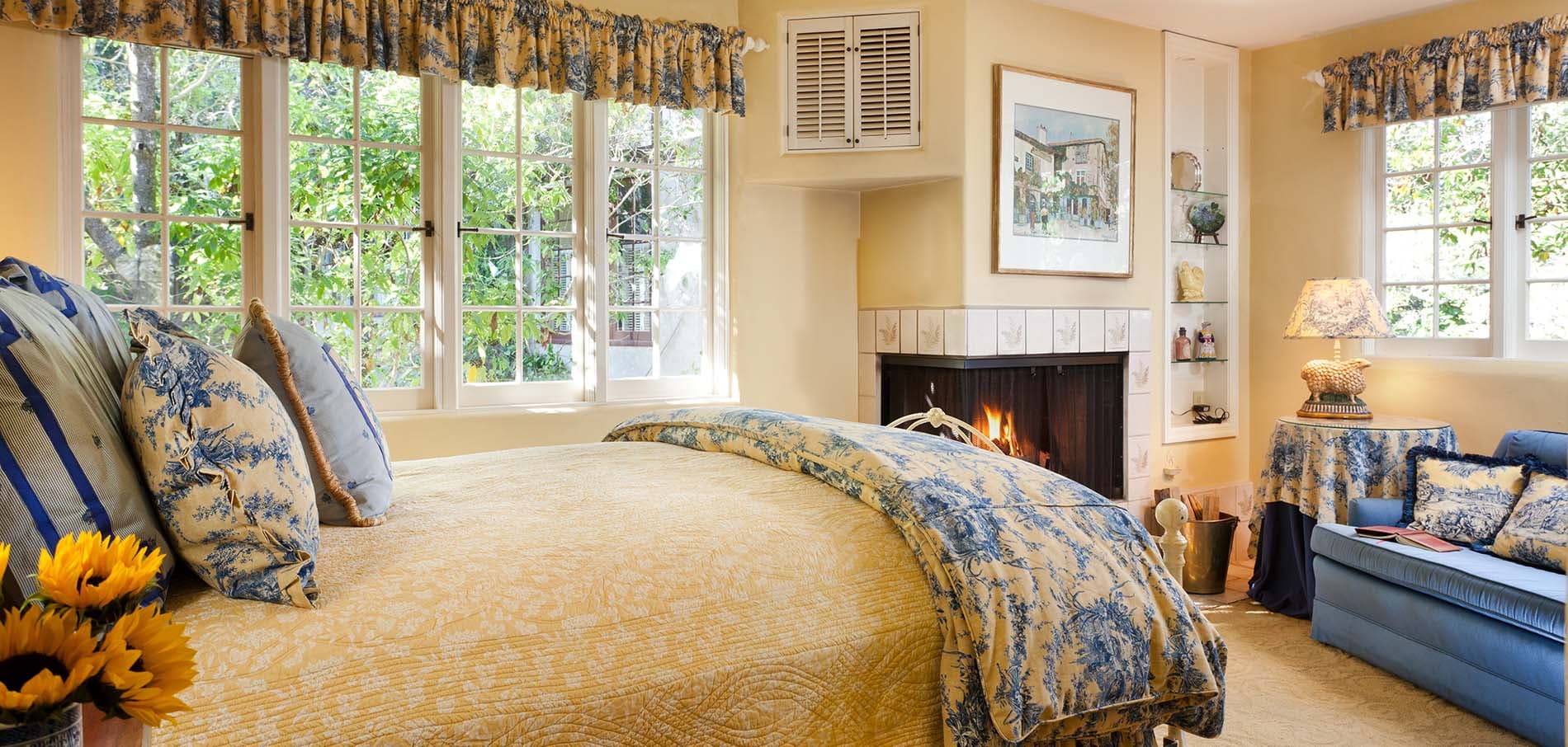 A cozy bedroom with a floral-patterned bedspread, large windows, and a fireplace.