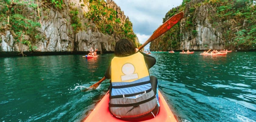 A person paddles a kayak through a narrow, rocky waterway surrounded by lush cliffs.