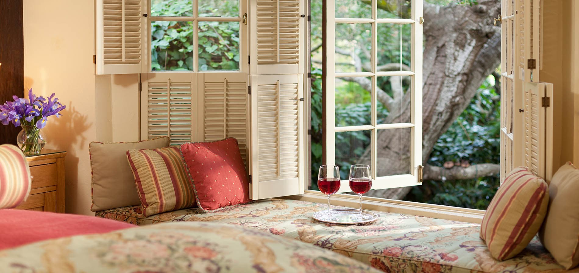 A cozy window seat with cushions and two glasses of red wine, overlooking a verdant garden.