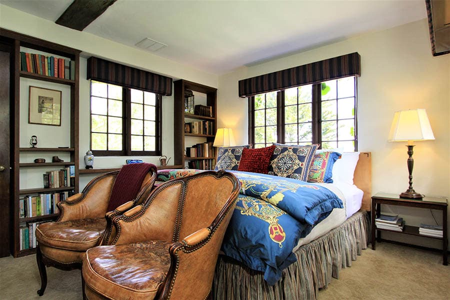 A cozy bedroom featuring a large bed with decorative pillows, two vintage leather chairs, and bookshelves.