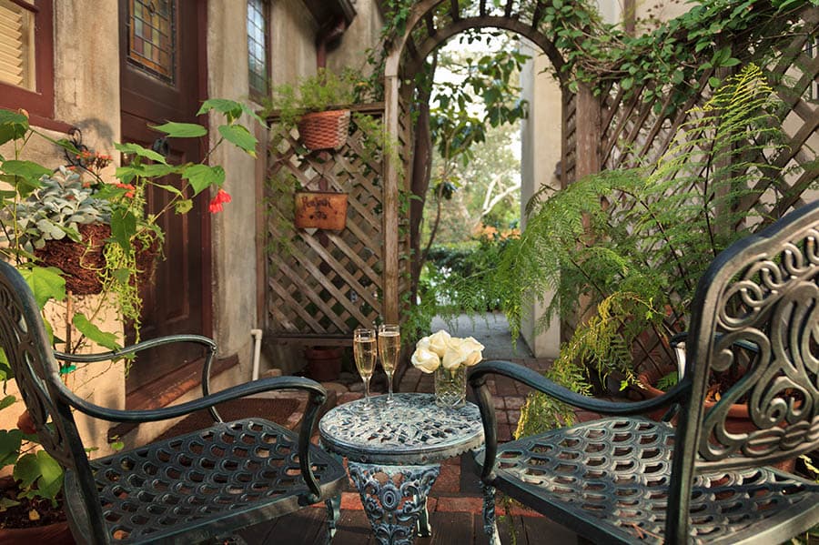 A cozy outdoor seating area with two chairs, a small table, champagne glasses, and white roses, surrounded by greenery and a lattice archway.