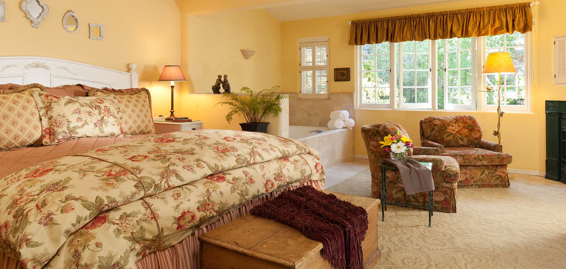 A cozy, yellow-hued bedroom featuring a floral-patterned bedspread, armchairs, and large windows.