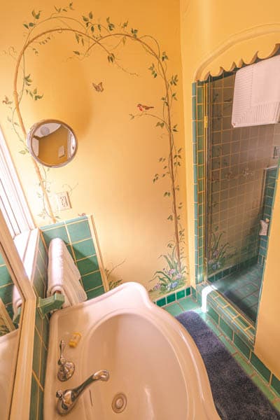 A vintage bathroom featuring a circular mirror, floral wall murals, a green tiled bathtub, and a separate shower area.
