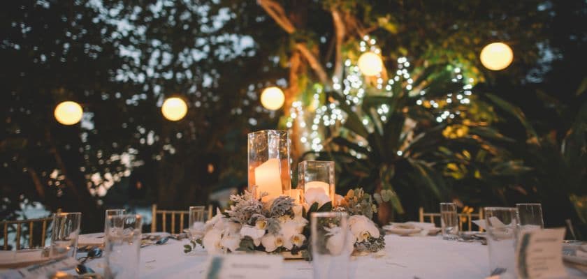 An elegant outdoor wedding table with candles, white flowers, glassware, and glowing lights in a garden setting.