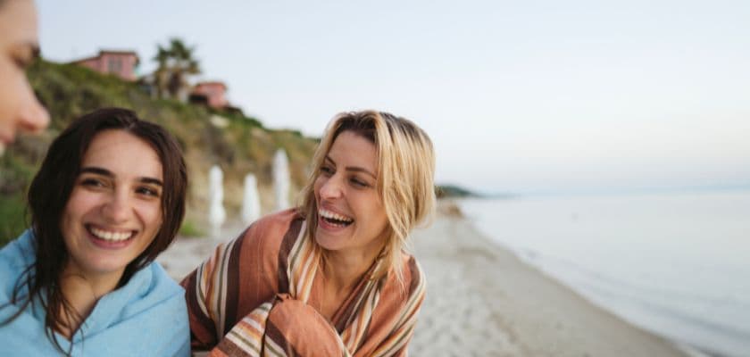 Women wrapped in striped blankets on a beach, looking and laughing at each other, with cliffs and calm water in the background.