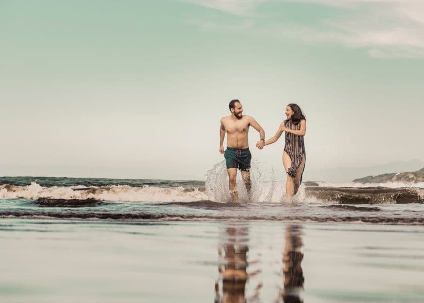 Couple holding hands and running through shallow ocean water with waves splashing around them under a clear sky
