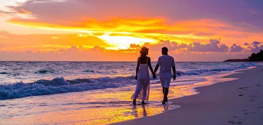 Couple walking hand in hand along the shoreline at sunset with waves rolling in and vibrant colors across the sky