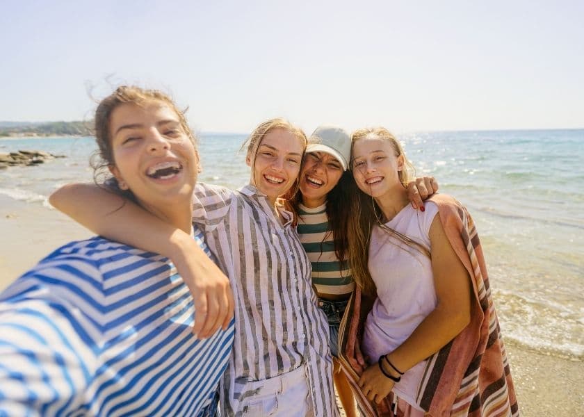 Four friends standing together on a beach near the water, posing for a selfie with the shoreline behind them.