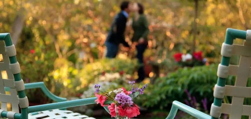 A romantic garden scene with flowers in the foreground and a couple sharing a kiss in the background.